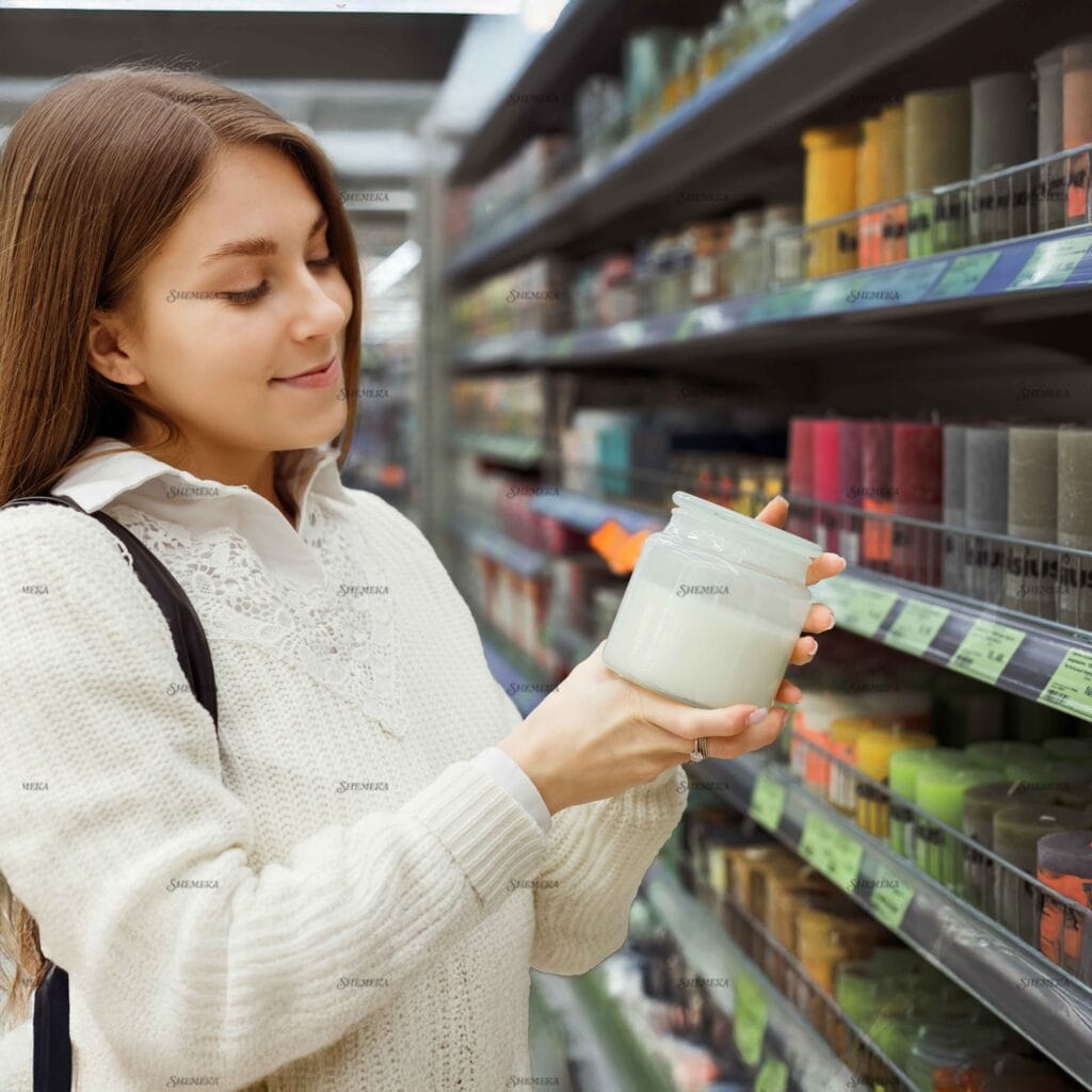 Young woman smelling scented candle in glass jar in shop.